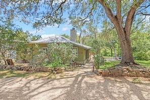 A rustic house with a chimney, surrounded by trees and a gravel path.