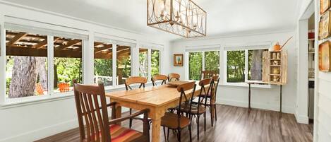 Un comedor con mesa y sillas de madera, ventanas amplias y una lámpara colgante.