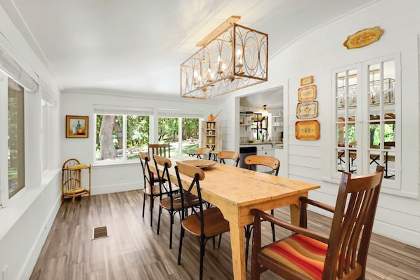 A dining area with a wooden table and chairs, a chandelier, and a view of the outdoors through large windows.