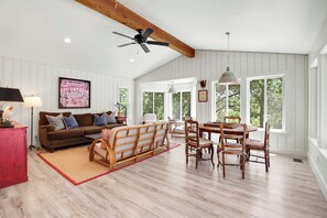 Un vaste salon doté d’un haut plafond et d’un ventilateur de plafond, un coin repas avec des chaises et une table en bois, un canapé marron agrémenté de coussins et de grandes fenêtres offrant une vue sur la verdure.