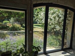 A room with a large window offering a view of a garden with white flowering trees and lavender plants. - Mas de la Gramillère (Barbentane)