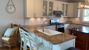 A kitchen with white cabinets, a granite countertop, a sink, a stove, and a dining area with chairs. - Tortuga: Spacious 5-bedroom home in Holden Beach, perfect for family fun! (Holden Beach)