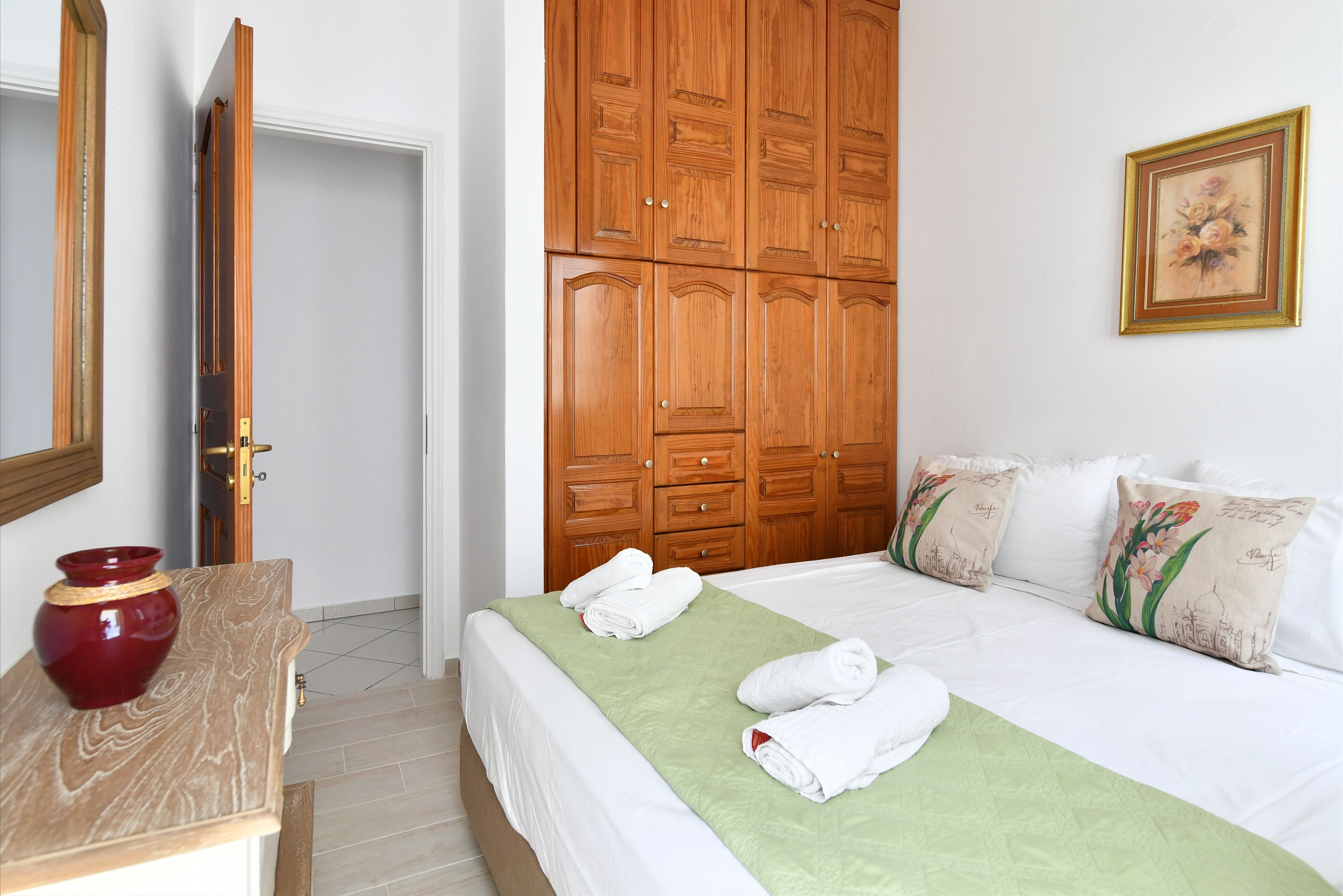 A hotel room with a bed, green bedspread, white pillows, a wooden wardrobe, and a red vase on a wooden table.