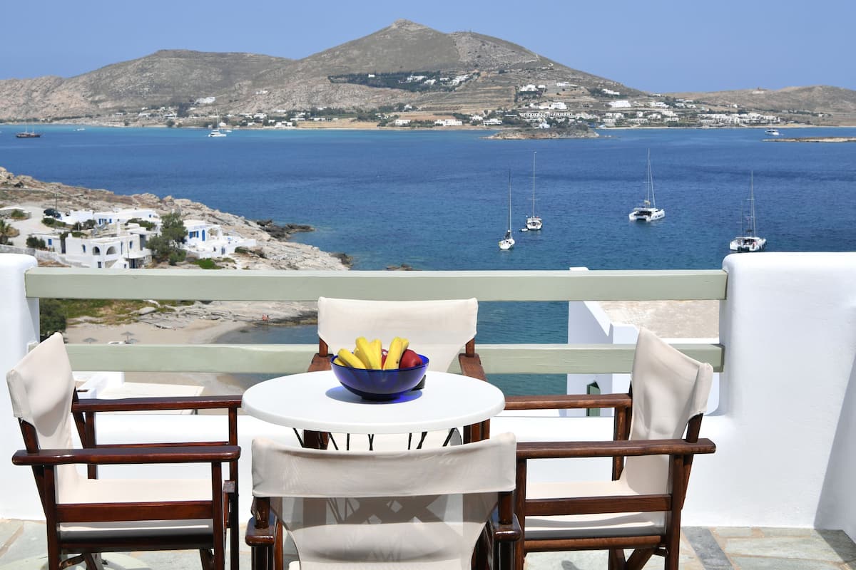 A balcony with two wooden chairs and a small round table, a bowl of fruit, and a view of the sea with boats and a mountain in the background.