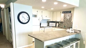A modern kitchen with white cabinets, a central island with bar stools, a clock, and a microwave.