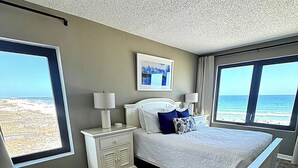 A bedroom with a large bed, nightstands, a framed picture, and a view of the beach.