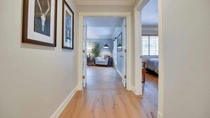 A hallway with wooden flooring, framed artwork on the walls, and a view into a bedroom with a bed and a lamp.