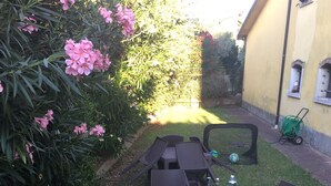 An outdoor dining area with a table set for two, surrounded by chairs, and a garden with pink flowers.