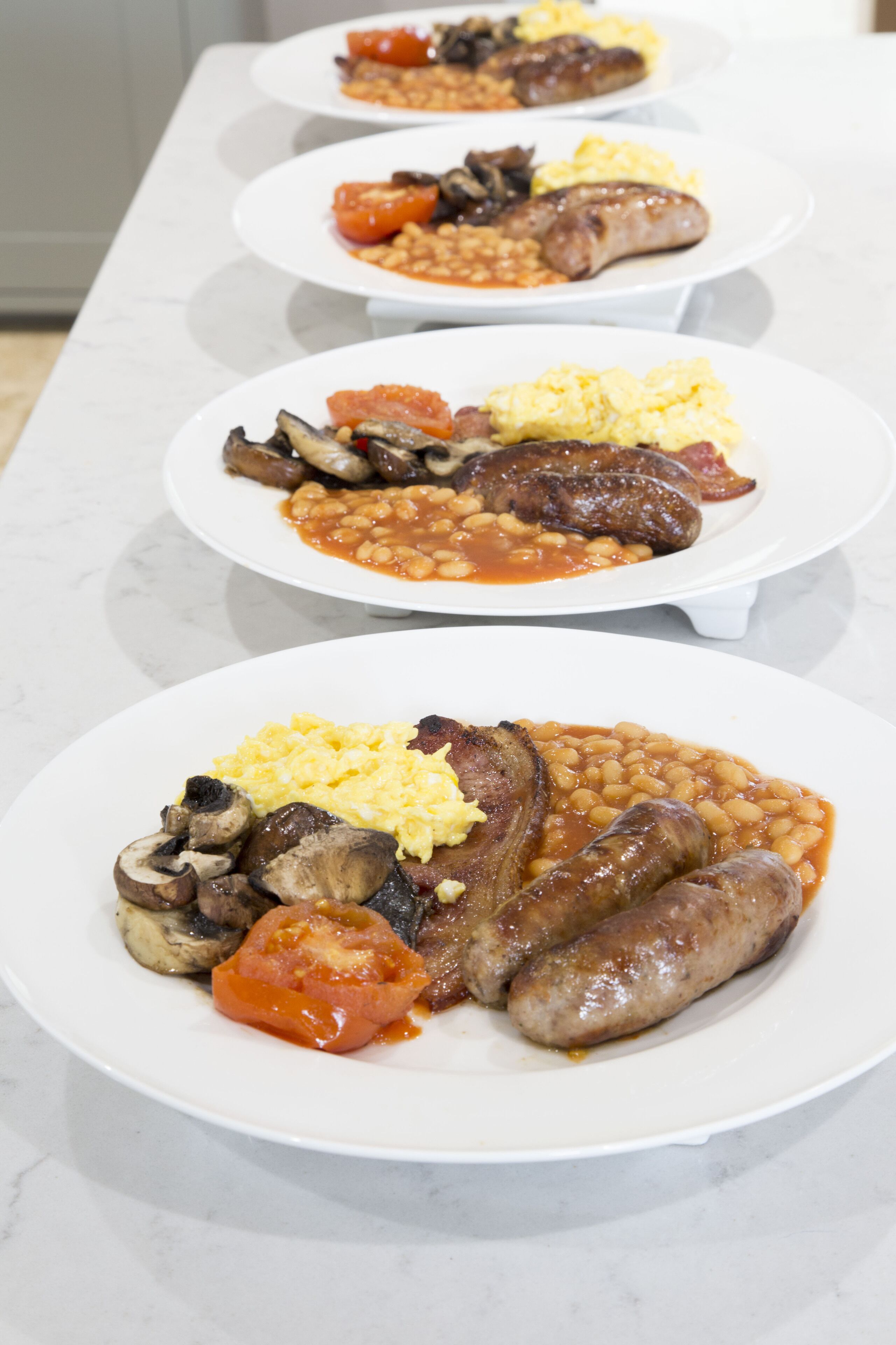 A row of plates with various breakfast items including sausages, beans, scrambled eggs, and tomatoes.