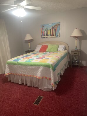 A bedroom with a bed, two bedside tables, a ceiling fan, a painting, and a red carpet. - Fishing/ Beach Getaway (Holden Beach)