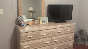 Une commode beige, surmontée d’un grand miroir, avec une télévision et divers objets de décoration.