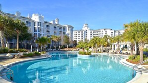 Una gran piscina al aire libre rodeada de palmeras y sillas de descanso, con un edificio de hotel de varios pisos al fondo.