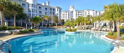 A large outdoor swimming pool surrounded by palm trees and lounge chairs, with a multi-story hotel building in the background.