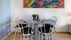 A dining area with a table set for four, white chairs with black cushions, a bottle of champagne, and two glasses. A colorful abstract painting hangs above the table. - Mediterraneo Seaview by Hello Homes Sitges (Sitges)