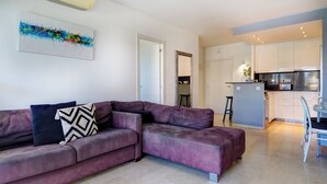 A modern living room with a purple sectional sofa, a dining area, and a kitchen with white cabinets. - Mediterraneo Seaview by Hello Homes Sitges (Sitges)