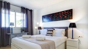 A modern bedroom with a large bed, bedside lamps, a nightstand, a window with curtains, and a painting on the wall. - Mediterraneo Seaview by Hello Homes Sitges (Sitges)