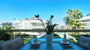 A balcony with a table set for two, a potted plant, and a view of a pool and buildings. - Mediterraneo Seaview by Hello Homes Sitges (Sitges)