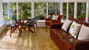 A living room with a glass table and chairs, a brown leather sofa with patterned cushions, and large windows offering a view of greenery. - Gardenside Lodge self - catering dog friendly holiday home in County Durham (County Durham)