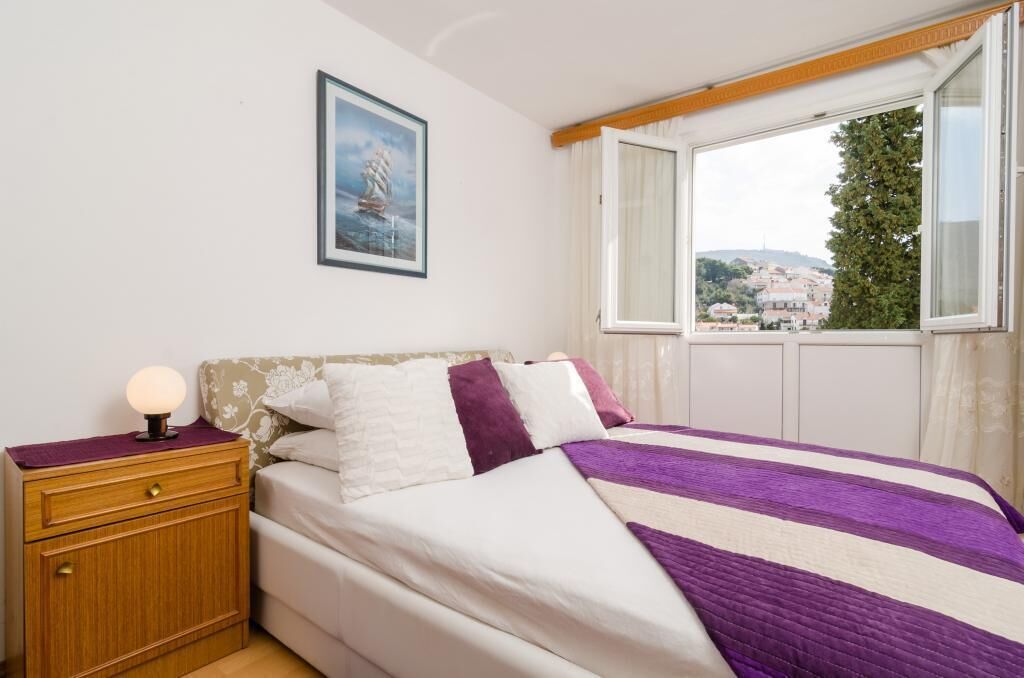 Une chambre à coucher comprenant un lit, une table de chevet, une lampe, une fenêtre donnant sur un arbre et des bâtiments, et un tableau encadré accroché au mur.