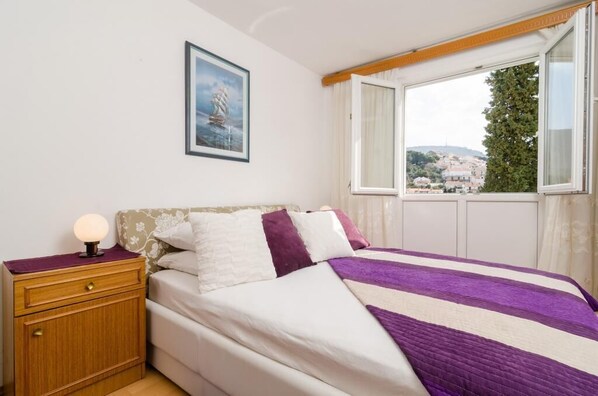 A bedroom with a bed, a nightstand, a lamp, a window with a view of a tree and buildings, and a framed picture on the wall.