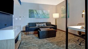 A modern hotel room with a grey sofa, a flat-screen TV, a desk with a lamp, and abstract wall art. - SpringHill Suites by Marriott Orangeburg (Orangeburg)