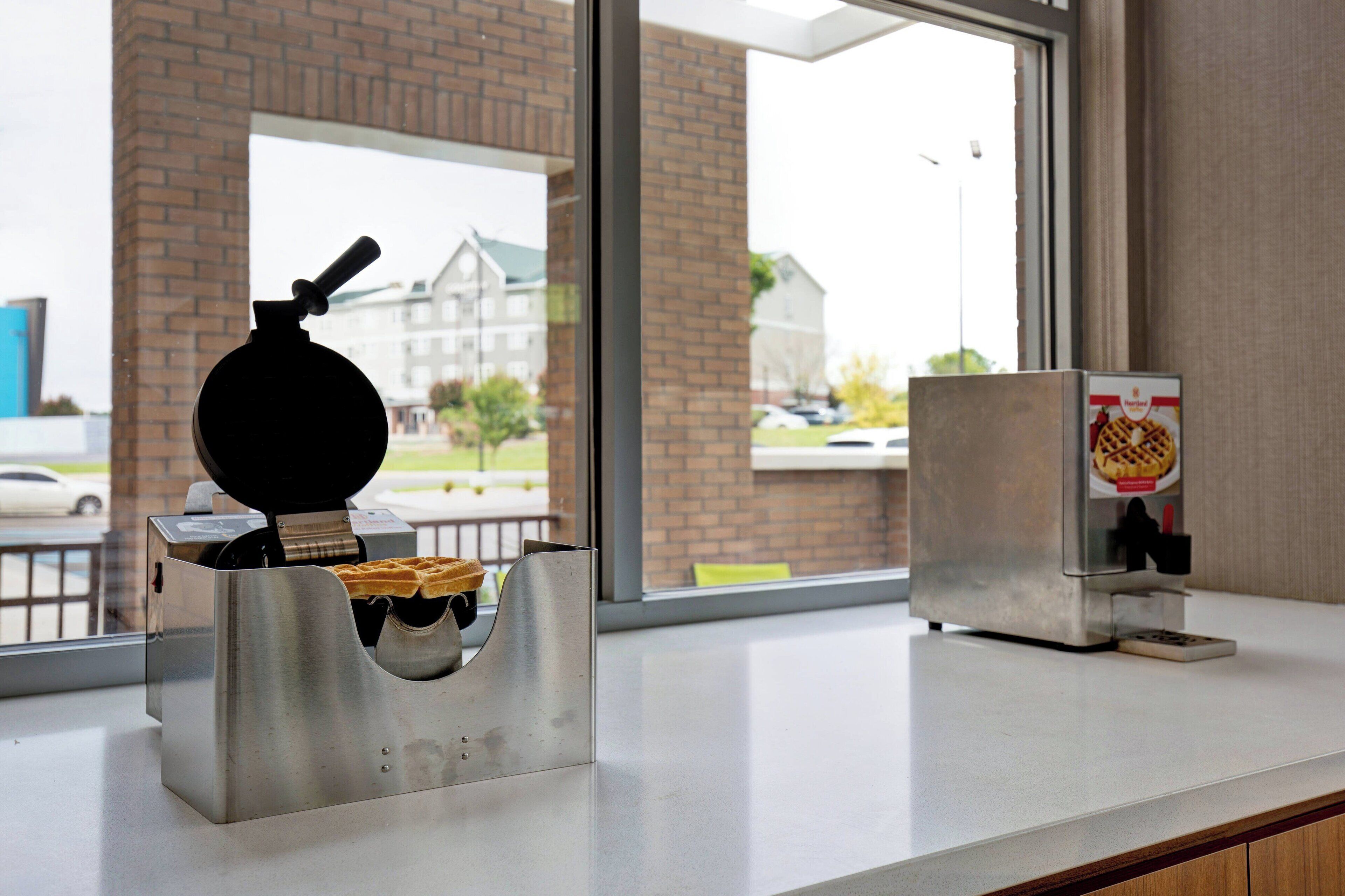 A waffle maker and a waffle dispenser on a counter with a view of buildings outside.