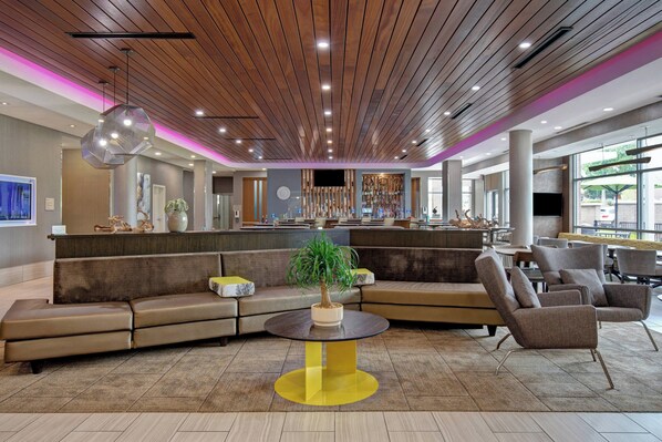 Um lobby moderno com teto de madeira, área de estar central com uma mesa redonda e um bar com variedade de garrafas.
