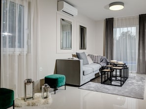 A modern living room with a sofa, a coffee table, and a side table. There is a window with curtains, an air conditioning unit, and a rug on the floor.