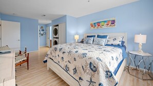 A bedroom with a large bed, a desk, and a chair. There is a door leading to another room and a wall-mounted artwork.