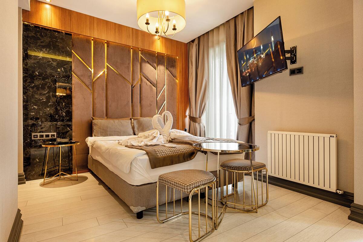 Una habitación de hotel moderna con una cama grande, una mesa de comedor redonda, dos taburetes, un televisor montado en la pared y una pared de acento de mármol.