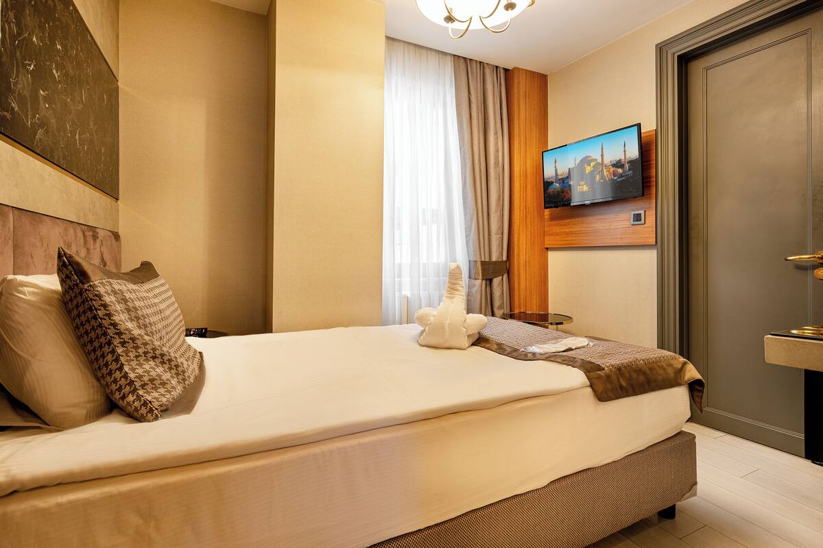 Una cama bien hecha con un almohadón, una mesita de noche con lámpara, un televisor montado en la pared y una puerta con picaporte.