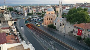 Eine Stadtansicht mit einer markanten Moschee, die ein großes Kuppelgewölbe und zwei Minarette zeigt, umgeben von modernen Gebäuden und einer belebten Straße mit Bussen und Autos.