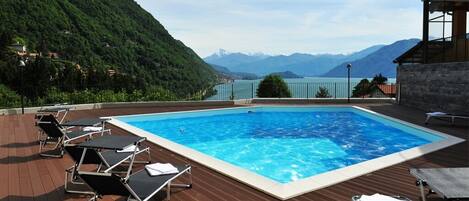 Piscina rettangolare circondata da lettini su un pontile in legno, con montagne sullo sfondo e cielo sereno.