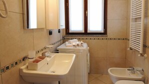 Un baño con lavamanos, bidé e inodoro. Hay una ventana con persianas, un secador de pelo y un espejo.