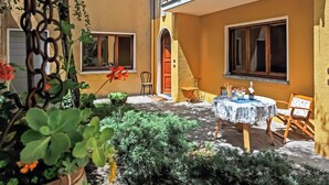 A courtyard with a table and chairs, surrounded by potted plants and a yellow building with large windows and an arched doorway. - Menaggio 57 (Menaggio)