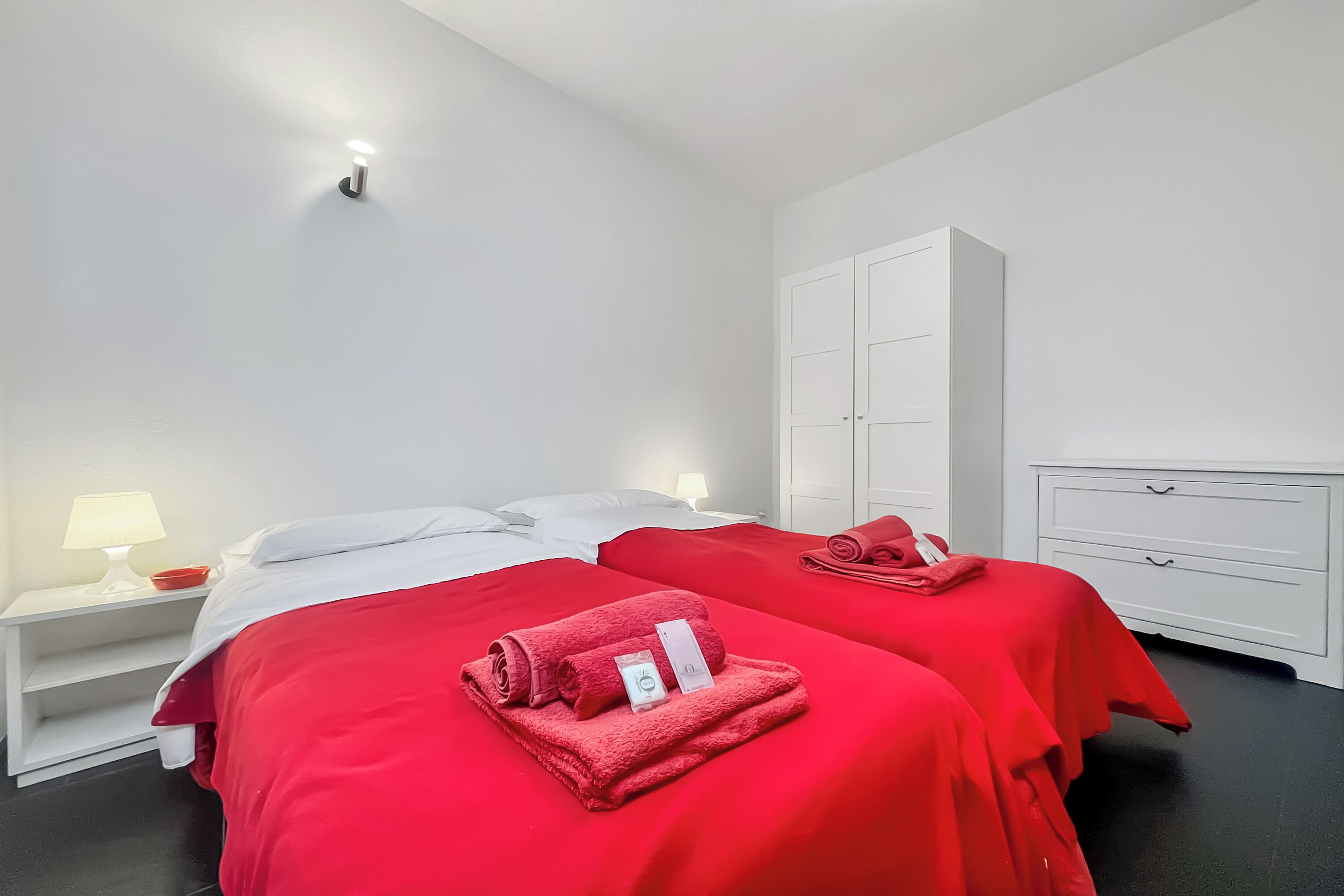 A hotel room with two beds, each with a red blanket and a rolled towel. There is a white wardrobe and a nightstand with a lamp.