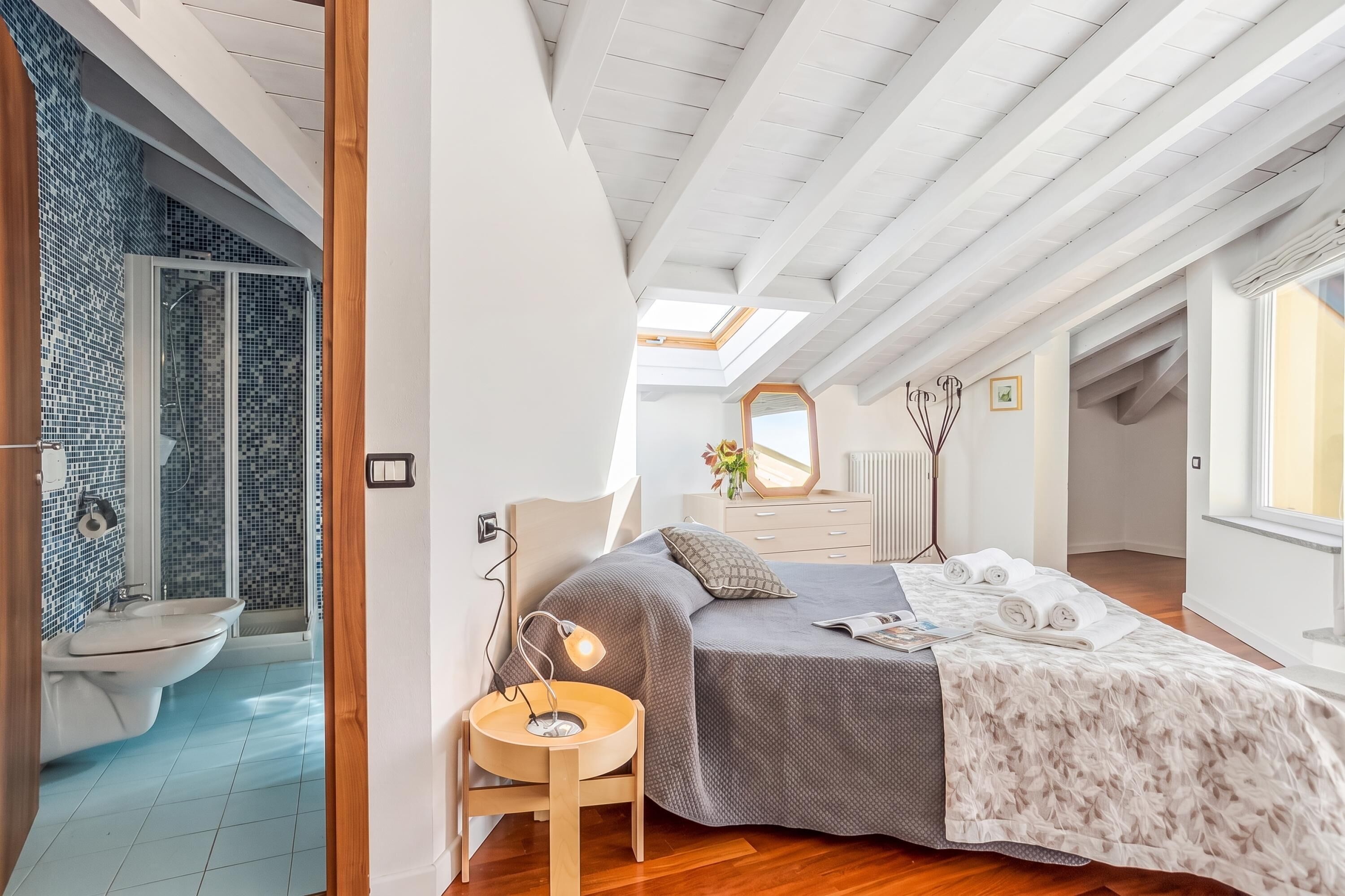 A bedroom with a skylight, a bed with a floral bedspread, a bedside table with a lamp, and a view of the bathroom.