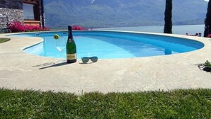 A swimming pool with a bottle of champagne and sunglasses on the edge, surrounded by a stone building and a mountain view. - Gera Panoramica (Gera Lario)
