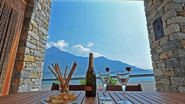 A table with a bottle of wine, two glasses, and a plate of chips, set against a stone wall with a view of mountains and a body of water. - Gera Panoramica (Gera Lario)