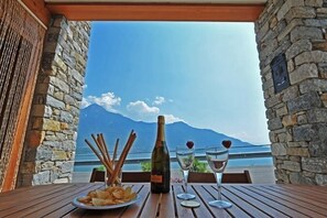 Una mesa con una botella de vino, dos copas y un plato de papas fritas, frente a un muro de piedra con vistas a montañas y un cuerpo de agua.