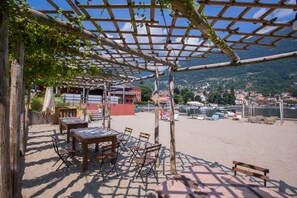 An outdoor seating area with tables and chairs under a wooden pergola, overlooking a waterfront with buildings and hills in the background.