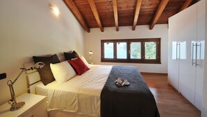 A bedroom with a wooden ceiling, a bed with white bedding and a black throw, a bedside table with a lamp, and a window with a view of greenery. - Bellano Perla Apt. 8 (Bellano)