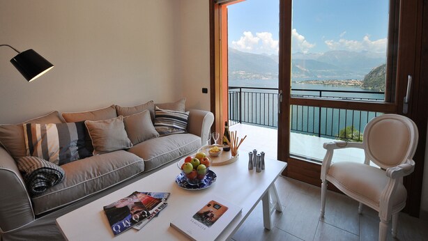 A modern living room with a sofa, a coffee table with magazines, a bowl of fruit, and a view of a lake and mountains through a large window. - Bellano Perla Apt. 8 (Bellano)