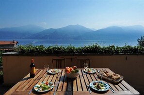 Una mesa de madera dispuesta con platos, vasos, una botella de vino y un bol de fruta, con vistas a un lago y montañas al fondo.