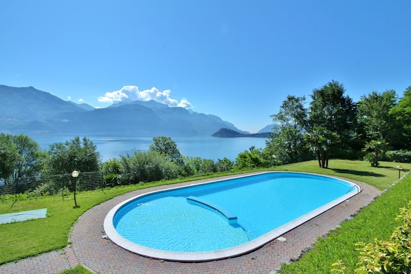 A swimming pool with a curved edge, surrounded by a brick pathway and lush greenery.