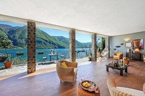 A spacious indoor area with a view of a lake and mountains, furnished with wicker chairs and a dining table. - La Torre Di Porlezza (Porlezza)