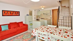 A compact living space with a red sofa, a dining table with a fruit-patterned tablecloth, a kitchen area, and a staircase. - Residenza Vercana Giardino (Vercana)