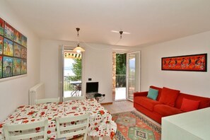 A room with a dining table, a red sofa, and a TV. There is a balcony with a view of greenery. - Residenza Vercana Giardino (Vercana)