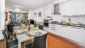 A modern kitchen with white cabinets, a dining area with a table set for four, and a large window with a view of a snowy landscape. - Bellavista Grande (Menaggio)