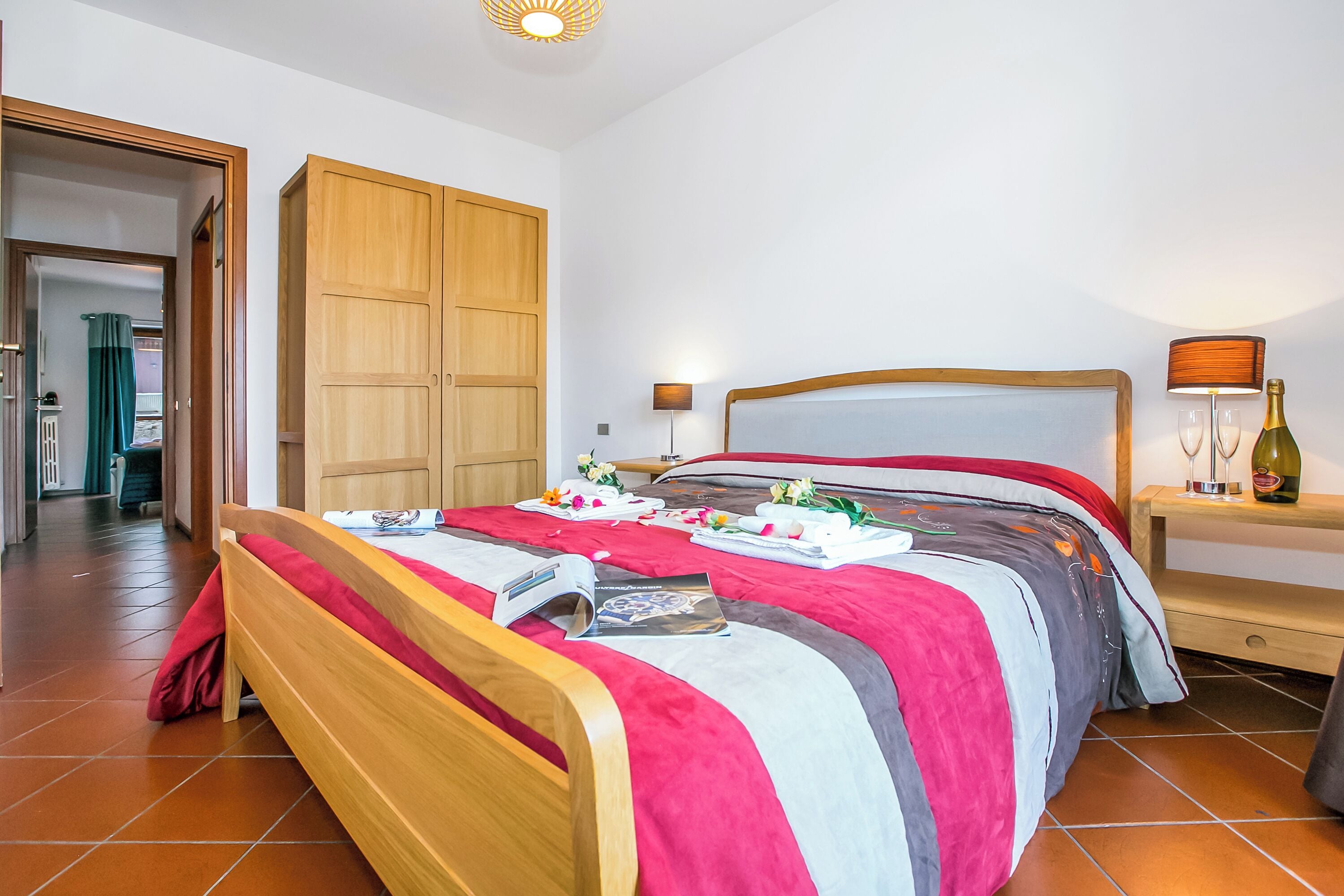 A neatly made bed with a red and grey bedspread, a wooden headboard, and a bedside table with a lamp and a bottle.
